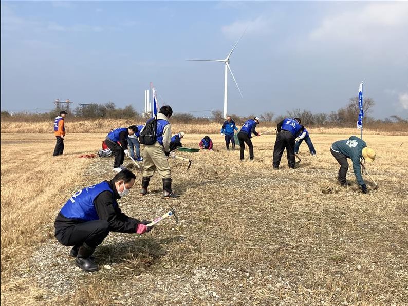 青いビブスを着た社員が風車の見える草地で除草作業をする様子