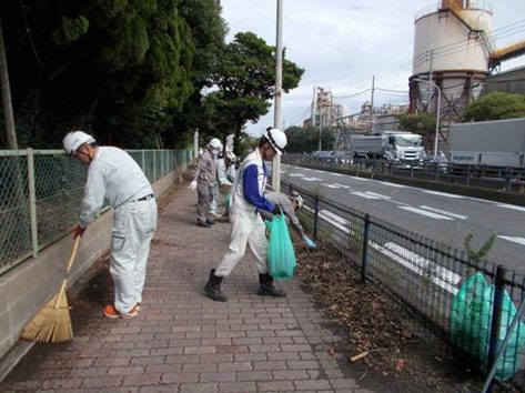 作業着姿の社員が歩道でほうきとごみ袋を手に清掃する様子